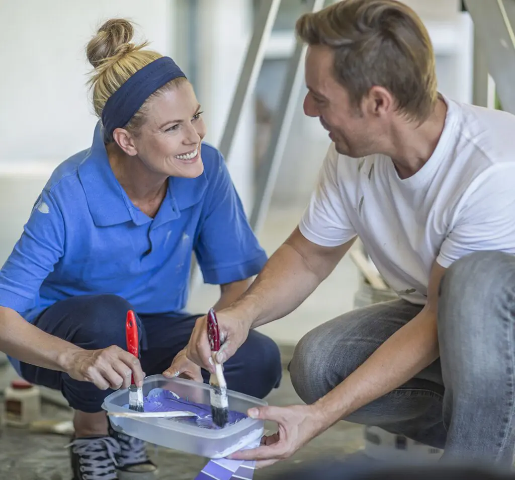 Couple crouching dipping paintbrushes in plastic container face to face smiling