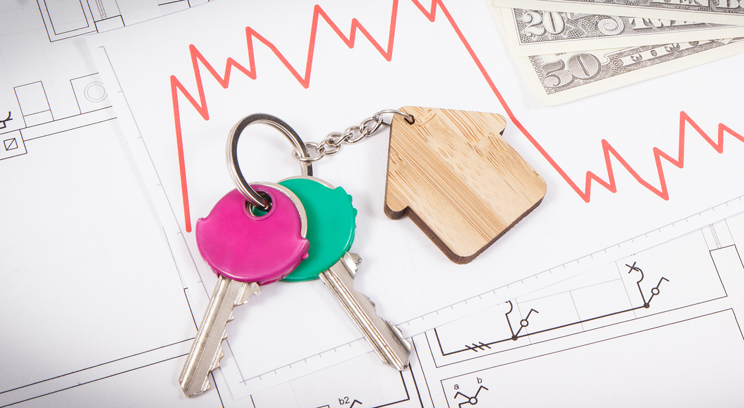 Home keys on a wooden house keychain sitting on top of graphs with money.